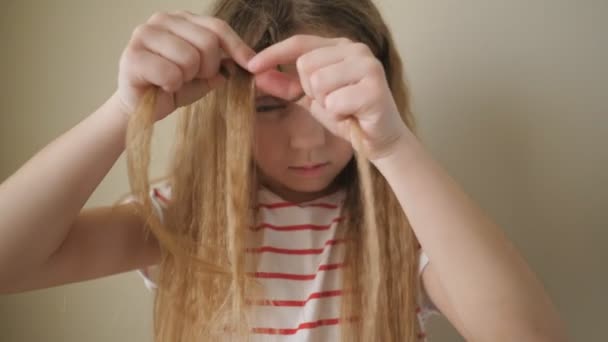 Zbliżenie na piękną małą dziewczynkę tkającą mały warkocz z jej długich blond włosów wewnątrz. Małe słodkie dziecko robi sobie fryzurę w domu. Zwolniony ruch — Wideo stockowe