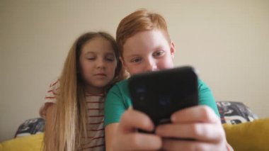 Gülümseyen küçük erkek ve kız kardeşlerin evde cep telefonuyla oyun oynamalarına yakın çekim. Küçük mutlu çocuk ve kız odasındaki kanepede oturmuş akıllı telefondan komik bir video izliyorlar. Dolly çekim ağır çekim
