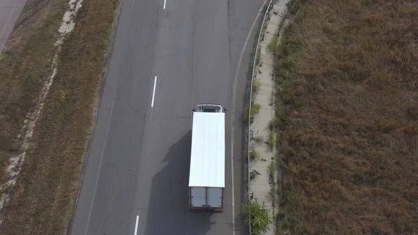 Aerial view of truck with white cargo trailer driving on road to destination. Flying over lorry moving through countryside highway. Concept of logistic or delivery. Top view Slow motion.