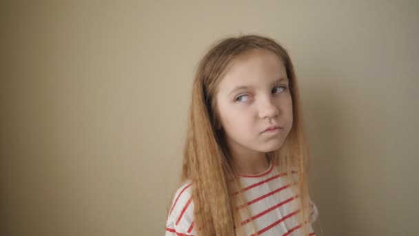 Niña cansada con el pelo largo y rubio posando a la cámara y balanceando su cuerpo de lado a lado en la habitación. Retrato de niño aburrido pequeño sobre el fondo de la pared beige. Primer plano: cámara lenta — Vídeo de stock