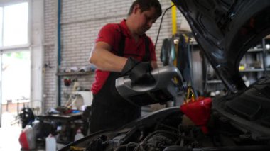 Siyah eldivenli tamirci, araçlar ve ekipmanlarla dolu profesyonel bir garaj ortamında araç bakımının ayrıntılı sürecini gösteren bir huniye motor yağı döküyor.