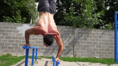 Sporcu paralel çubuklar üzerinde dinamik amuda kaldırma eğitimi veriyor, arka planda canlı yeşillikle ilerleyen bir çalışma sekansında güç ve dengeyi gösteriyor.