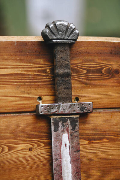 Hilt and part of the sword blade, half pushed out of the sheath, close-up against a blurred wooden wall.
