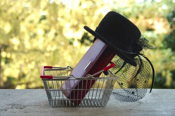 Shopping basket with red purse and small black female hat with veil and feather in retro style. Shopping cart with gift or present on bokeh effect background. Black Friday and Shopping sale concept.