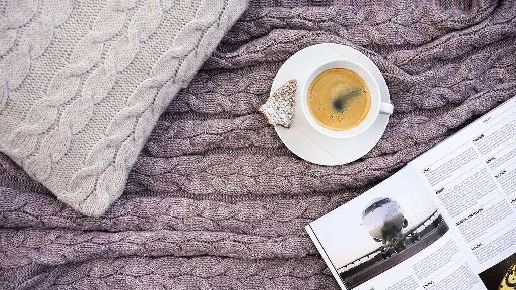 White cup of hot aromatic coffee with belgium waffle cookie in the form of fir and white pages of book for traveling on the gray pillow and warm woolen gray pink plaid. Knitted textile with a braid