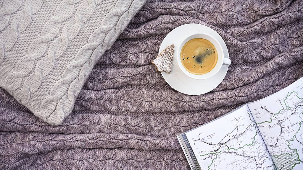 White cup coffee with flock of birds in sky and waffle cookie in form fir, white pages book with map traveling on pillow and warm woolen gray plaid. Winter autumn concept comfort and dreams.