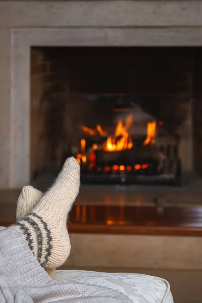 Warm white knitted socks on the feet of unrecognizable woman or man sitting with cozy plaid in front of the fireplace. Cozy relaxed magical atmosphere home interior. Christmas New Year winter concept.