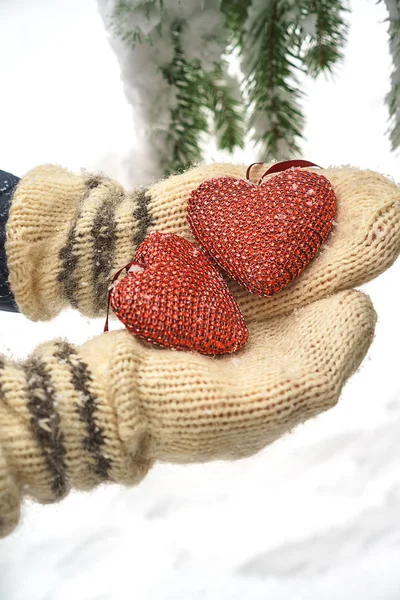 Dwa czerwone włókienniczych serca i ręce na tle oddział ciężkich ośnieżone jodły, w pobliżu Dom z czerwonej cegły. Wesołych Świąt, szczęśliwego nowego roku i Walentynki pozdrowienie składu, Koral kolor serca — Zdjęcie stockowe