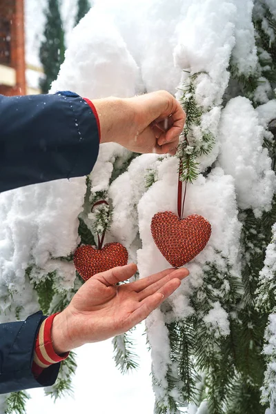 Dwa czerwone włókienniczych serca i mans ręce na tle oddział ciężkich ośnieżone jodły, w pobliżu Dom z czerwonej cegły. Wesołych Świąt, szczęśliwego nowego roku i Walentynki pozdrowienie składu, Koral kolor serca — Zdjęcie stockowe