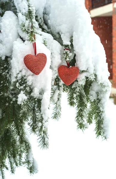 Dwa serca czerwone tkaniny wisi na oddziale ciężkich ośnieżone jodły, w pobliżu Dom z czerwonej cegły. Wesołych Świąt, szczęśliwego nowego roku i Walentynki koncepcja pozdrowienie — Zdjęcie stockowe