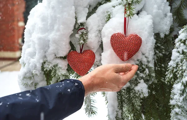 Dwa czerwone włókienniczych serca i mans ręce na tle oddział ciężkich ośnieżone jodły, w pobliżu Dom z czerwonej cegły. Wesołych Świąt, szczęśliwego nowego roku i Walentynki pozdrowienie składu, Koral kolor serca — Zdjęcie stockowe