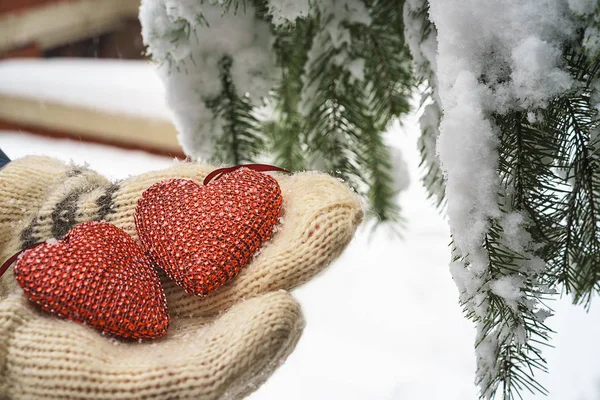 Two red textile hearts and hands on heavy snowy fir branch background, near red brick house. Merry Christmas, Happy New Year and Valentines Day greeting composition, coral color of heart
