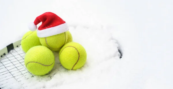 Santa hat on tennis ball, set of tennis balls on racket on white snow winter background. Merry Christmas and New year concept with tennis balls play. Close up, sport lifestyle, funny.