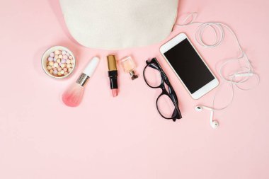 Flat lay female accessories: cosmetics, glasses, mobile phone, headphones, makeup on pastel pink background.