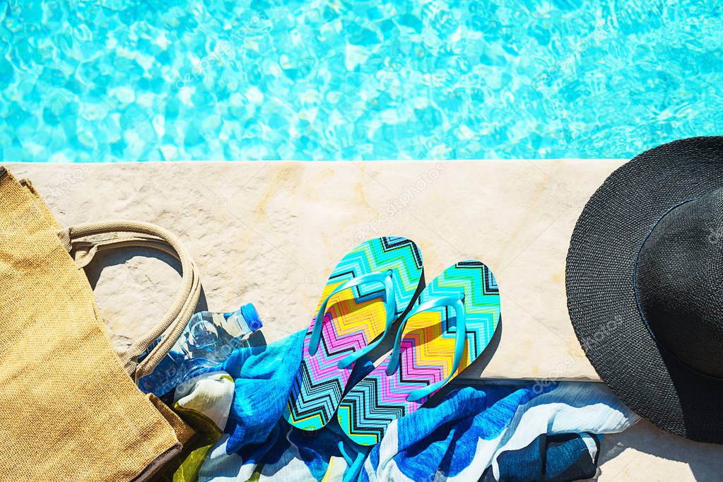 Swimming pool and straw hat, colorful flip flops, wicker bag, straw hat, bottle with still water as sunny summer background.