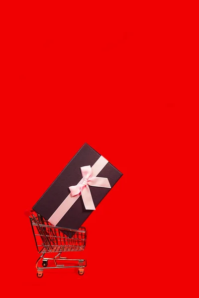 Regalo de vacaciones, concepto de Viernes Negro con caja de regalo y lazo rosa en un carrito de compras de metal, canasta sobre fondo de color rojo . — Foto de Stock