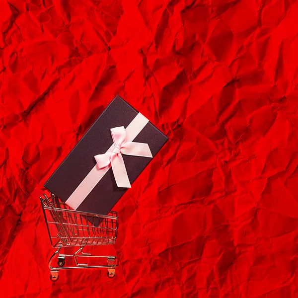 Regalo navideño, concepto de Viernes Negro con caja de regalo y lazo rosa en un carrito de compras de metal, canasta en papel de envolver de color rojo . — Foto de Stock