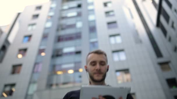 ヘッドフォンを持ち タブレットを手に持った男がビジネスセンターの近くのタブレットで入力している — ストック動画