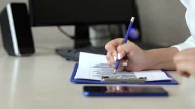 İş kadını bir ofis masasında otururken iş çizelgeleriyle çalışıyor. Yanında bir cep telefonu var.