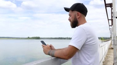 Genç bir adam elinde telefonla köprüde duruyor.