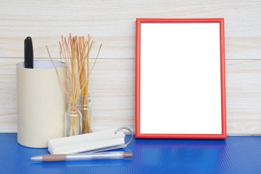 Beyaz boşluklu kırmızı fotoğraf çerçevesi, ahşap duvar zeminine karşı mavi masaya yerleştirilmiş. Silindirik kutu, cam şişedeki kurumuş dallar, yaratıcı sunum için notlar ve kalemle şekillendirilmiş.                               
