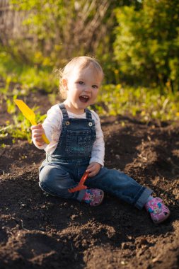 Yerde kürekle oturan bebek ve güneş ışığında komisyon. Sevimli yaz sarışın kız bahçede