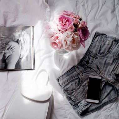 Düz lay, dergiler, sosyal medya. Üstten görünüm. Güzellik blog kavramı. Kadın moda aksesuarları, şort, buket gül ve pions, telefon ve kahve beyaz yatak zemin üzerine