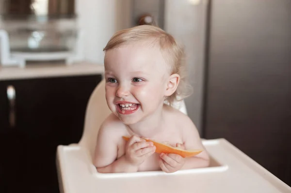 Bebé comiendo melón melón. Gracioso niño riendo en la cocina. Alegre niño en la silla alta —  Fotos de Stock