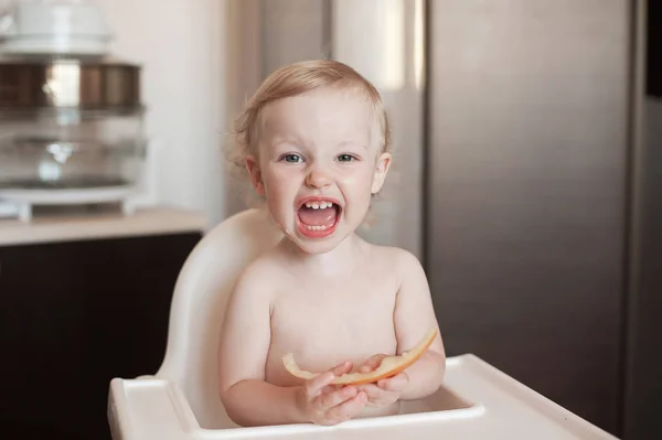 Μωρό κορίτσι τρώει πεπόνι μελιτώματος. Αστείο γέλιο παιδιού στην κουζίνα. Χαρούμενο παιδί στο καρεκλάκι — Φωτογραφία Αρχείου