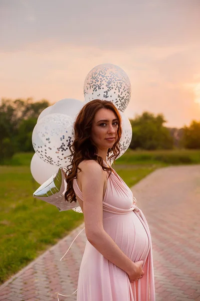 Feliz bela mulher grávida em vestido longo rosa andando com balões de ar no parque verde ao nascer do sol dia de verão . — Fotografia de Stock