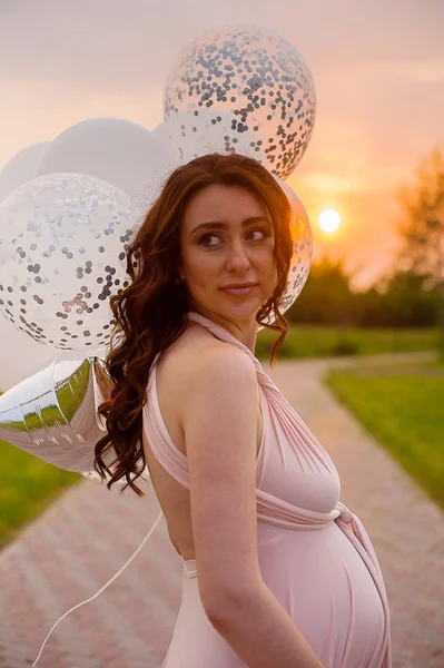 Feliz bela mulher grávida em vestido longo rosa andando com balões de ar no parque verde ao nascer do sol dia de verão . — Fotografia de Stock