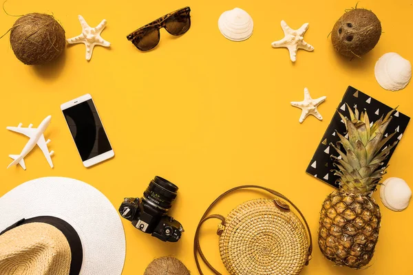 Summer traveler accessories flat lay. Straw hat, retro film camera, bamboo bag, sunglasses, coconut, pineapple, sea shells and starfish, air plane, notebook and phone over yellow background
