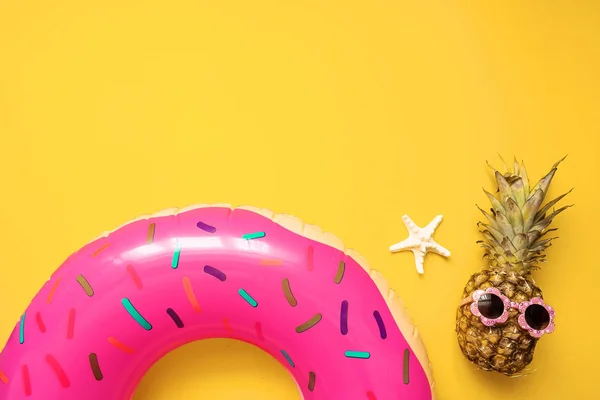 Colorful summer flat lay with pink inflatable circle donut, funny pineapple in sunglasses and starfish starfish over yellow background, top view. Holiday concept