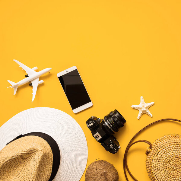 Summer traveler accessories flat lay. Straw hat, retro film camera, bamboo bag, sunglasses, coconut, pineapple, starfish, air plane, notebook and phone over yellow background, top view.