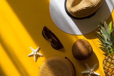 Renkli yaz kadın moda kıyafeti düz yerde yatıyordu. Hasır şapka, bambu çanta, güneş gözlüğü, hindistan cevizi, ananas, sarı arka planda deniz yıldızı, üst manzara. Yaz modası, tatil konsepti