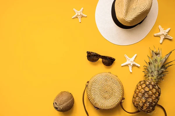 Colorful summer female fashion outfit flat lay. Straw hat, bamboo bag, sunglasses, coconut, pineapple and starfish over yellow background, top view. Summer fashion, holiday concept. — Stock Photo, Image