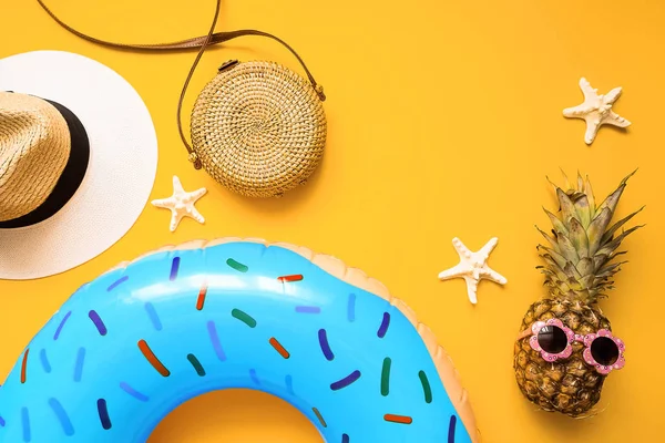 Colorful summer flat lay with blue inflatable circle donut, funny pineapple in sunglasses, straw hat, bamboo bag and starfish starfish over yellow background, top view. Holiday concept