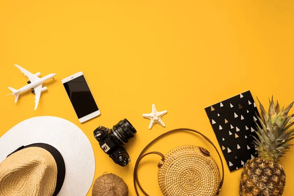 Summer traveler accessories flat lay. Straw hat, retro film camera, bamboo bag, sunglasses, coconut, pineapple, sea shells and starfish, air plane, notebook and phone over yellow background