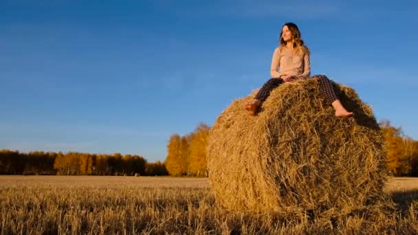Laughing young woman throws shoes at the camera on haystacks. Pretty girl in casual clothes gets fun on farmer field — Stock Video