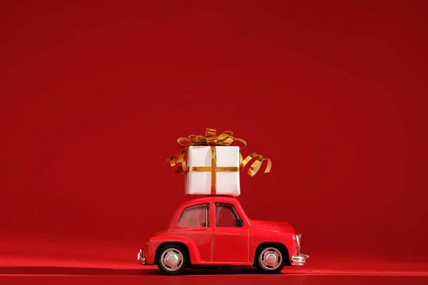 Red small retro toy car with gift on the roof on red background. Delivery. New Year, Christmas, Valentines Day, World Womans Day, Sale concept.