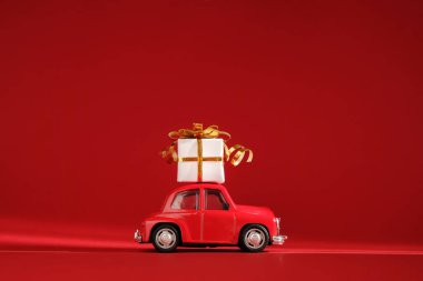 Kırmızı arka planda çatıda hediyeli küçük kırmızı oyuncak araba. Teslimat. Yeni Yıl, Noel, Sevgililer Günü, Dünya Kadın Günü, İndirim Konsepti.