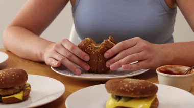 Aşırı yemek. Bir kadın hamburger yer, diğer hamburgercilerle dolu tabaklar yakınlarda durur. Bir sürü ev yapımı hamburger. Yürekli bir öğle yemeği. Bir kız çok hamburger yer, eller yakın plan. Aşırı yeme sorunu kavramı.