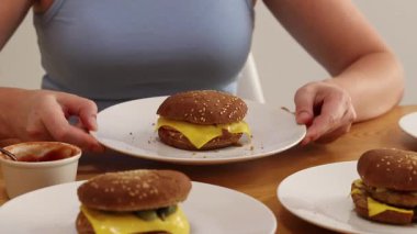 Aşırı yemek. Bir kadın hamburger yer, diğer hamburgercilerle dolu tabaklar yakınlarda durur. Bir sürü ev yapımı hamburger. Yürekli bir öğle yemeği. Bir kız çok hamburger yer, eller yakın plan. Aşırı yeme sorunu kavramı.