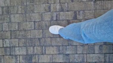 Beyaz spor ayakkabılar ve kaldırımlı taş patika boyunca yürüyen kot pantolonlu kadın bacakları. Yaz yürüyüşü. Doğa yürüyüşü. Parkta yürüyen bacaklar. Üst Manzara. Açık hava yürüyüşü