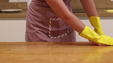 Cleaning lady wipes kitchen table. Woman in apron and yellow gloves wipes table with viscose napkin. Cleaning apartment from dust and dirt. Cleaning service. Close-up of hands in cleaning gloves