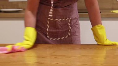 Cleaning lady wipes kitchen table. Woman in apron and yellow gloves wipes table with viscose napkin. Cleaning apartment from dust and dirt. Cleaning service. Close-up of hands in cleaning gloves