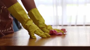 Cleaning lady wipes kitchen table. Woman in apron and yellow gloves wipes table with viscose napkin. Cleaning apartment from dust and dirt. Cleaning service. Close-up of hands in cleaning gloves