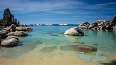 Lake Tahoe North, Nevada county, California, ABD'de Kum Limanı'nın uzun pozlama, birkaç bulutlar ile mavi bir gökyüzü gününde arka plan dağların tepesinde mavi şeffaf su, kayalık kıyı ve kar featuring- seyahat Kaliforniya
