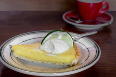 Bir bar ın ahşap tezgahında kırmızı fincan kahve bir fincan ile beyaz tabakta krem şanti ile tepesinde anahtar kireç pasta bir dilim