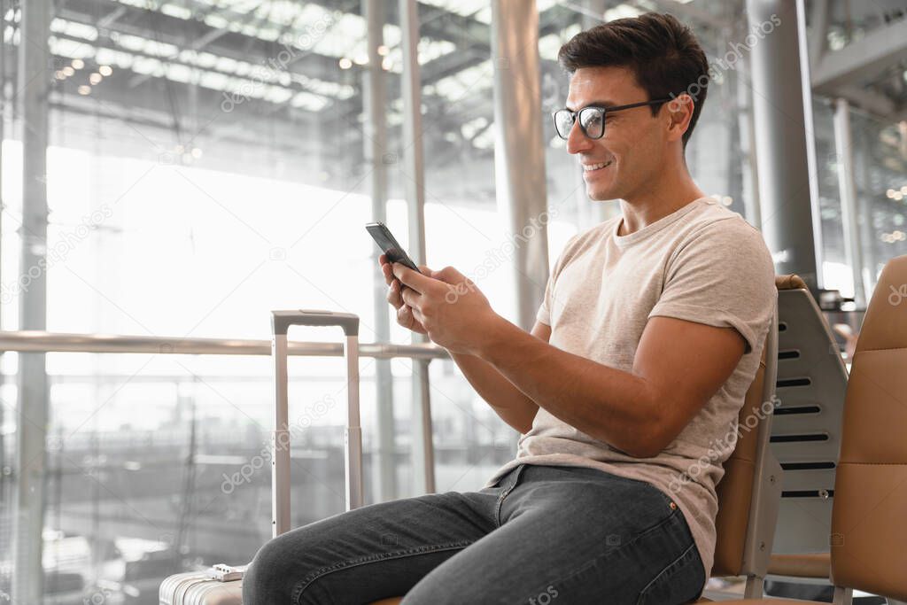 Tourist man relaxing look at his cell phone and smiling happily at the airport terminal waiting for boarding with copy space, traveling protection, concept of travel insurance, holiday alone.